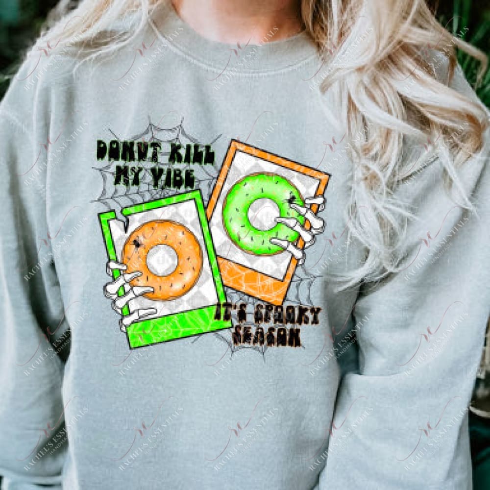 Blonde model wearing a grey crewneck sweatshirt with a halloween design. The design says Donut kill my vibe, It's spooky season. The design has to polaroid picture frames, neon green & orange with 2 sprinkled donuts. Skeleton hands are holding the picture frames and spider webs and spiders are behind the wording