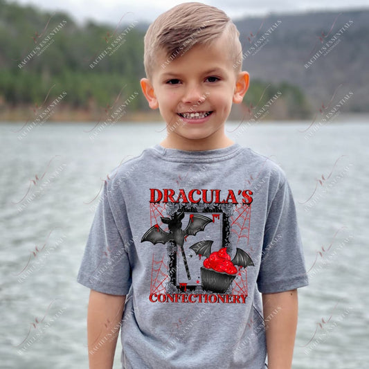 child male model wearing a grey short sleeve t-shirt featuring a design that says Drucula's Confectionery. The design is an antique black picture frame with red spider webs surrounding it. Inside the frame is a black cupcake with red frosting and bat wings emerging from it. There is a also a bat candy on a stick.