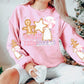 Model wearing a sweatshirt featuring a Christmas design on the front and on both sleeves. The design on the front says North Pole Cookie Co. and features a gingerbread man, a peppermint and gingerbread cookies shaped like a star and christmas tree. The sleeve is the same design without the wording.