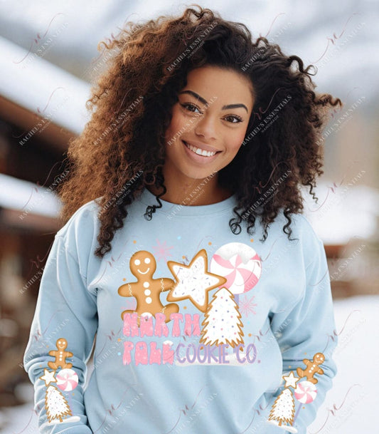 Model wearing a sweatshirt featuring a Christmas design on the front and on both sleeves. The design on the front says North Pole Cookie Co. and features a gingerbread man, a peppermint and gingerbread cookies shaped like a star and christmas tree. The sleeve is the same design without the wording.