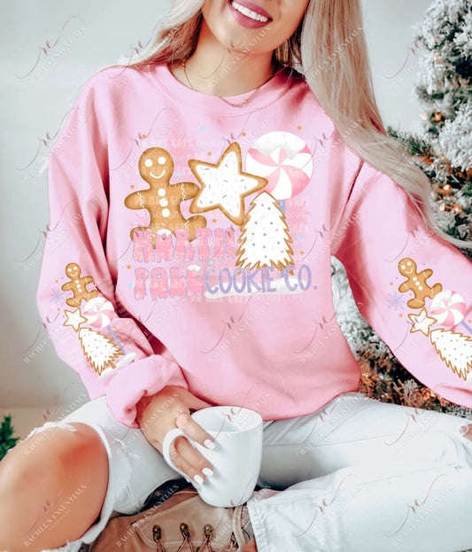 Model wearing a sweatshirt featuring a Christmas design on the front and on both sleeves. The design on the front says North Pole Cookie Co. and features a gingerbread man, a peppermint and gingerbread cookies shaped like a star and christmas tree. The sleeve is the same design without the wording.