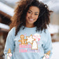 Model wearing a sweatshirt featuring a Christmas design on the front and on both sleeves. The design on the front says North Pole Cookie Co. and features a gingerbread man, a peppermint and gingerbread cookies shaped like a star and christmas tree. The sleeve is the same design without the wording.