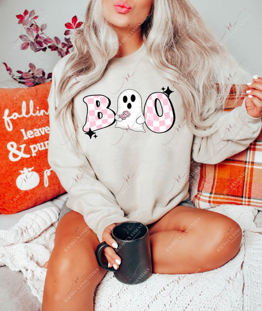 Blonde model wearing a tan colored sweatshirt featuring the word boo in pink checkered lettering. Instead of the middle o, a white ghost wearing a pink ring pop is featured. Black stars are on the outer design