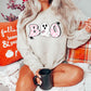 Blonde model wearing a tan colored sweatshirt featuring the word boo in pink checkered lettering. Instead of the middle o, a white ghost wearing a pink ring pop is featured. Black stars are on the outer design