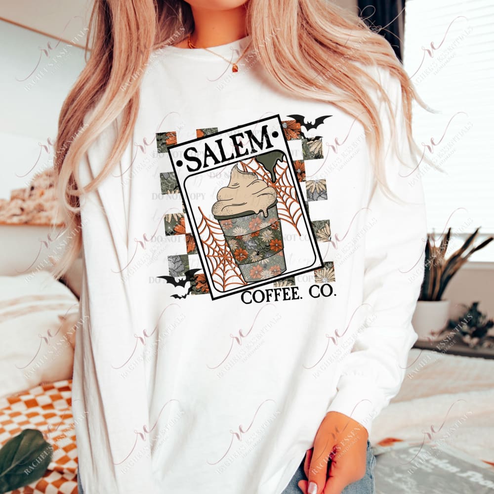 A model wearing a white sweatshirt featuring a halloween design. A checkered distressed orange and yellow floral background. In the front is a sign that signs Salem Coffee Co. with a latte with whip cream and orange spider webs