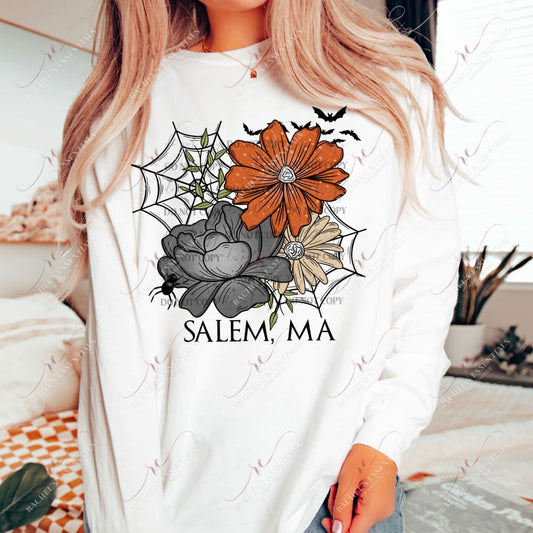 Model wearing a white sweatshirt featuring a halloween design. The design has 2 spiderwbs in the background and 3 large flowers in orange, black and yellow. with the wording Salem, MA at the bottom. Bats are flying at the top of the design and a spider is crawling on the black flower