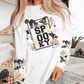 Model wearing a wearing a white long sleeve shirt with a halloween design on the front and on the sleeves. The front shows an image with the words spooky and a black cat, witches hat, ghosts, bats, spiders and mushrooms surrounding it. The sleeve features the same design without the wording. 