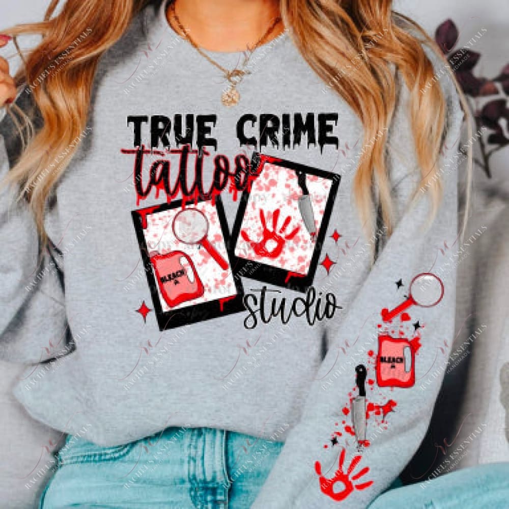 Model wearing a grey sweatshirt with a true crime tattoo studio. The design features two pictures with a red bleach bottle and magnifying glass in one and a bloody hand print and knife in the other. Blood splatter, a hand print, bleach, and magnifying glass are featured on the sleeve on the sweatshirt. 